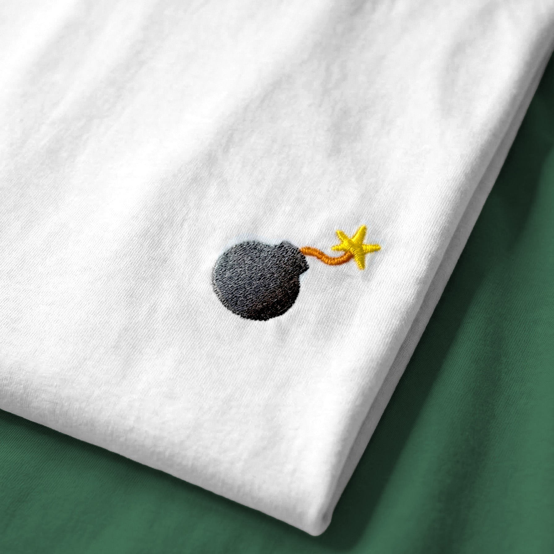 White t-shirt with a small embroidered design of a bomb on a green surface