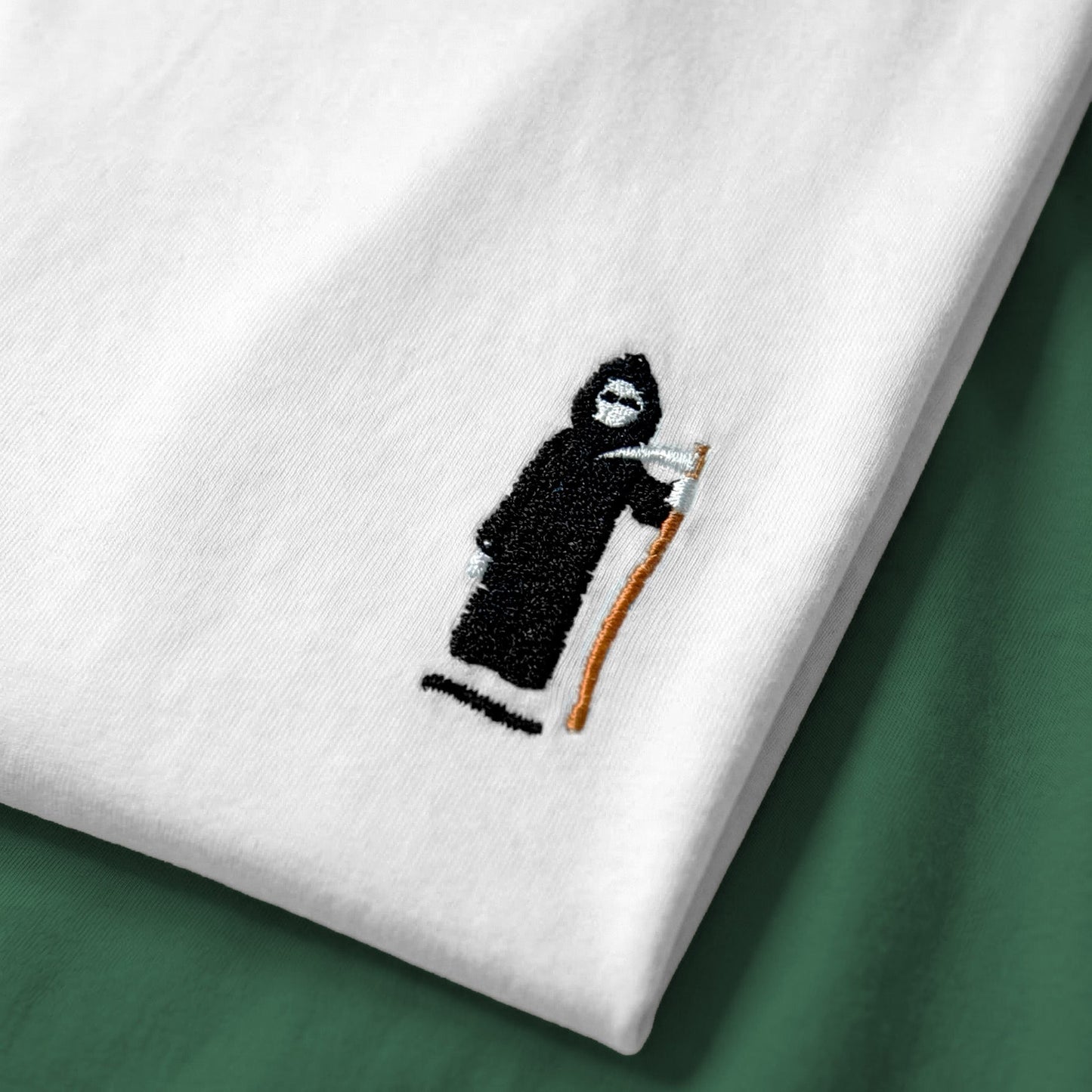 White tee with a grim reaper embroidered design on a green surface